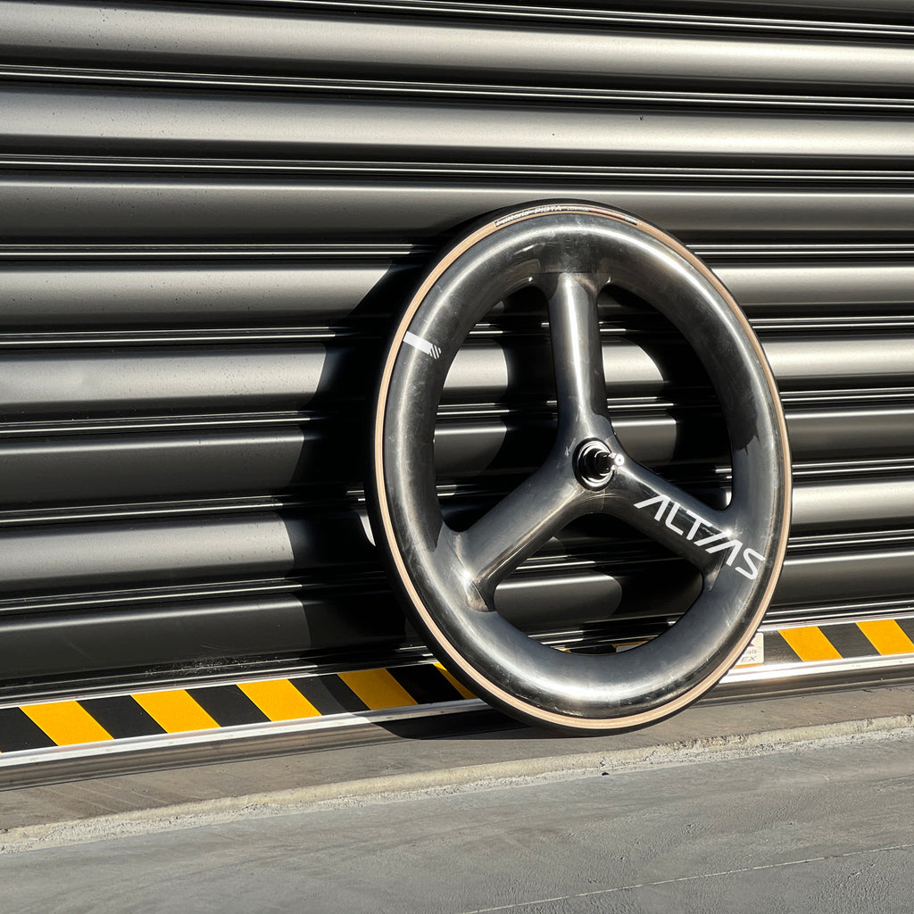 A carbon fiber bicycle wheel with a dark gray finish and white 'ALTIAS' branding, leaning against a dark gray metal roller shutter.