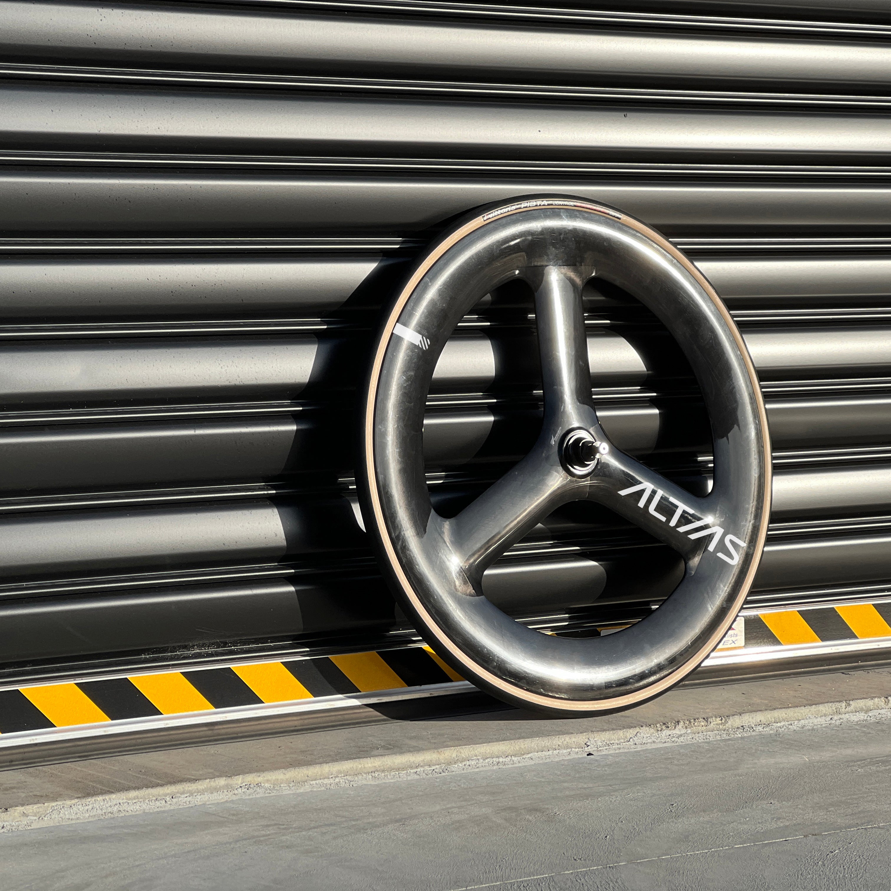 A carbon fiber bicycle wheel with a dark gray finish and white 'ALTIAS' branding, leaning against a dark gray metal roller shutter.