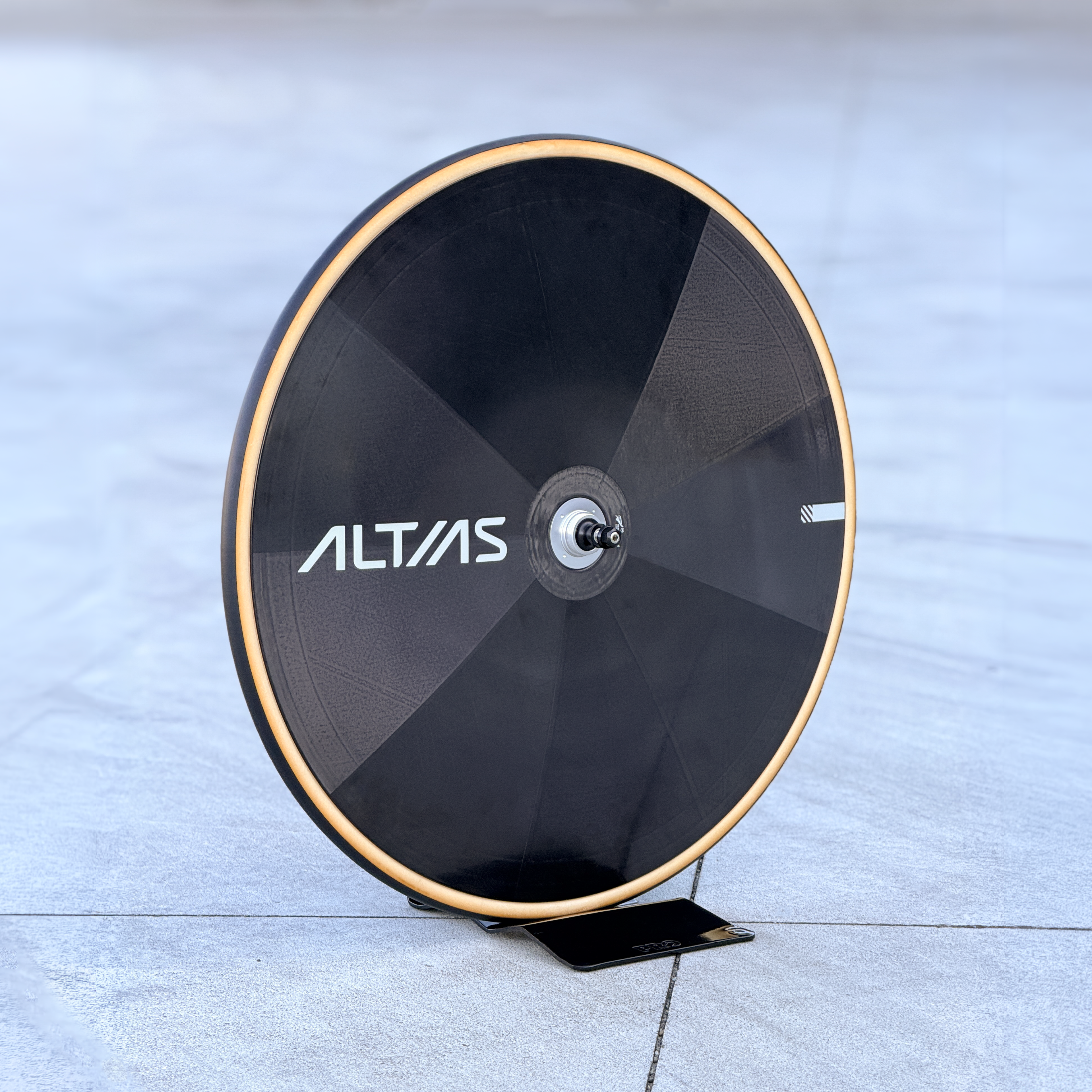 A black Altas disc wheek with a tan coloured tyre, displayed on a small black stand on a gray concrete floor.
