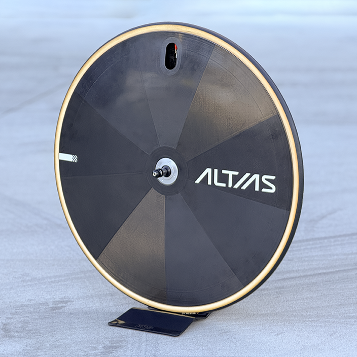 A black Altas disc wheel bicycle component with a tan-colored tyre, standing on a small black support against a light gray concrete background.
