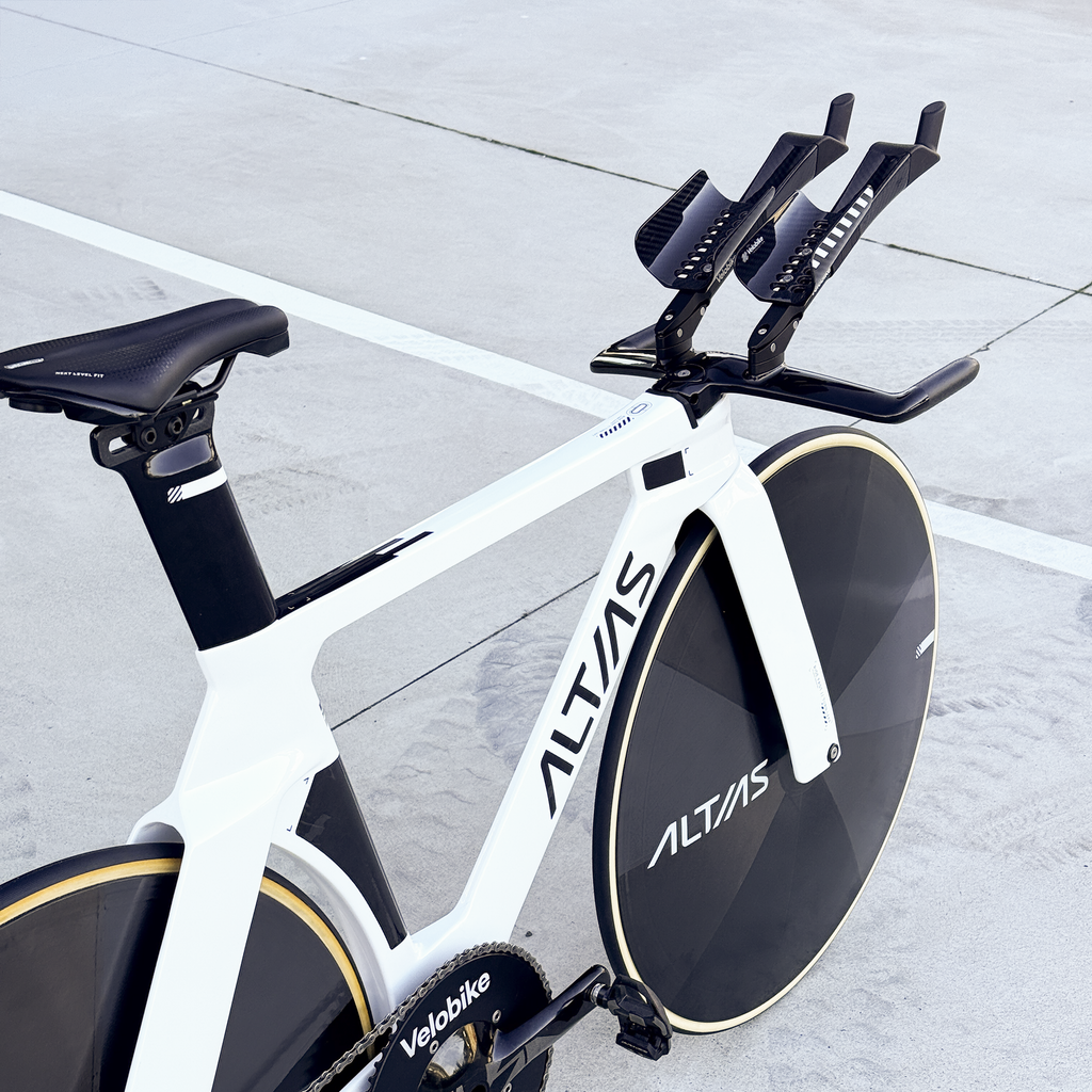 A white and black Altas track bicycle with a large black disc wheel, black handlebars, and a black saddle, photographed on a light concrete surface with white painted lines.