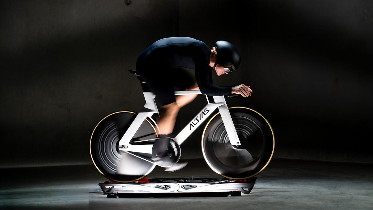 A cyclist in a black aerodynamic suit and helmet rides a white ALVAS time-trial bike on a stationary trainer in a dark studio setting.