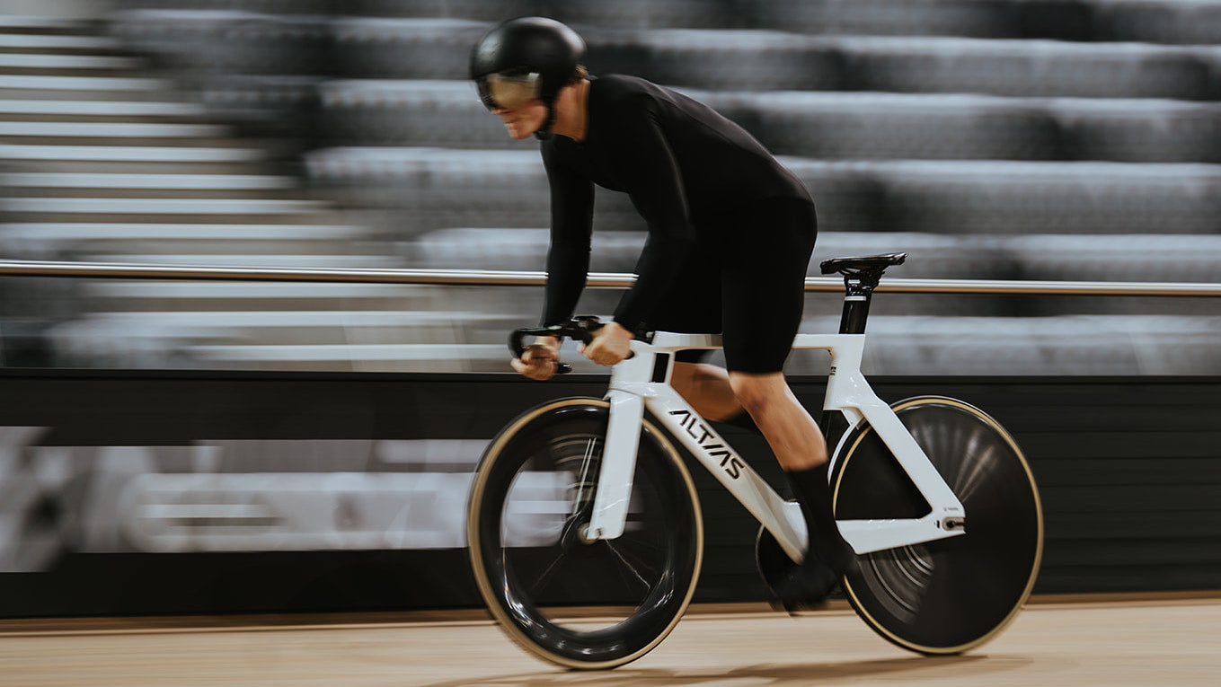 A cyclist in a black outfit rides a white and black Alzans track bicycle on an indoor velodrome track with blurred stadium seating in the background.
